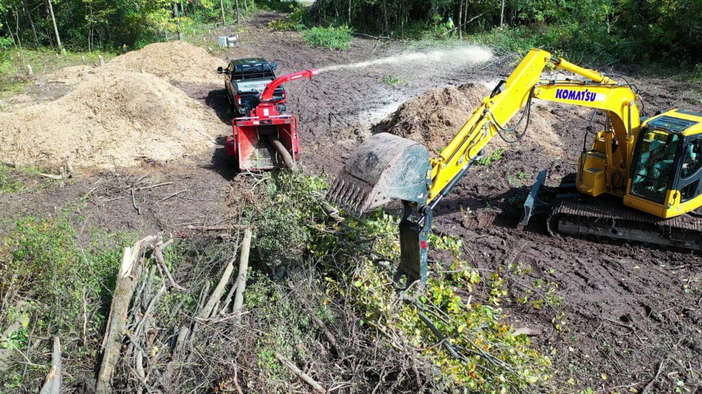 Aerial view of a Komatsu excavator with a grapple attachment feeding brush into a red wood chipper on a land clearing site.