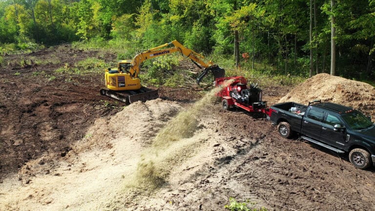 A Komatsu excavator feeding brush into a wood chipper during a large-scale forestry mulching and land clearing project.