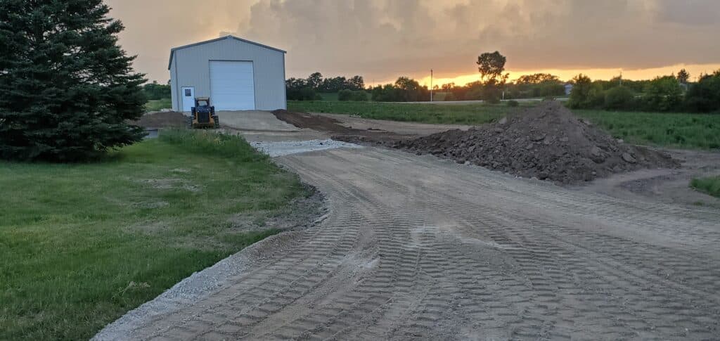 A professionally graded gravel driveway sub-base leading to a large metal pole barn at sunset.