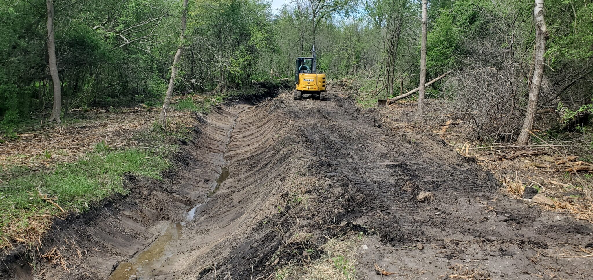 Excavation Services A John Deere excavator carving out a precise drainage swale through a wooded area to manage water runoff.